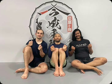 Shaker Zaman seated on the mat with Masakazu Imanari and a training partner under the Imanari Jiu-Jitsu logo
